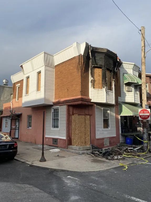 Corner house with fire damage around the upper bay window, with plywood covering the front door and the windows.
