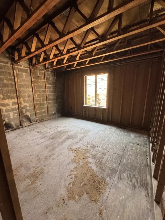 The room stripped to bare wall framing, a block wall, exposed ceiling joists, and a plywood subfloor.