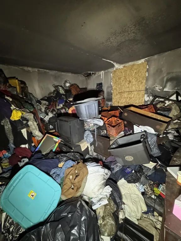 Room with smoke blackened walls and ceiling, filled with piled clothing, bins, and household items, and a boarded window.