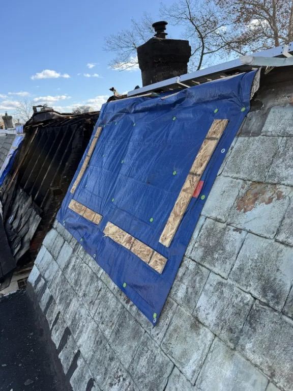 Blue tarp fastened with wood strips over a section of a slate roof, beside a charred section of roof and a chimney.