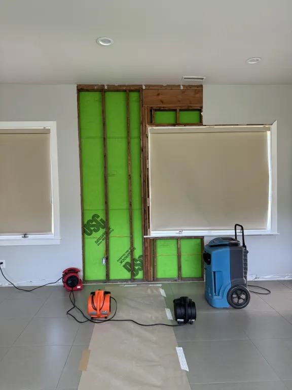 Wall opened to the framing beside a window, with a blue dehumidifier and three air movers on the floor.