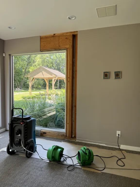 Room with the wall opened beside and above a large window, with a dehumidifier and two green air movers on floor protection.